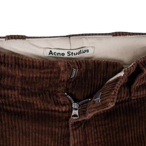 May include: Close-up of brown corduroy pants with a zipper and button closure. The pants have a visible label that reads "Acne Studios". The corduroy fabric has a ribbed texture.