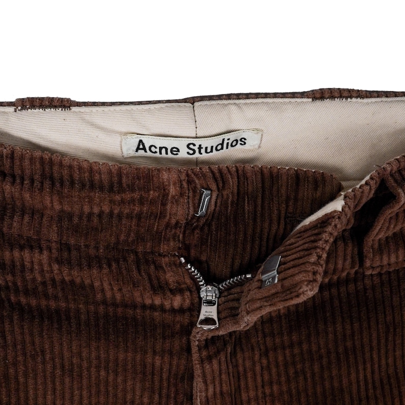 May include: Close-up of brown corduroy pants with a zipper and button closure. The pants have a visible label that reads "Acne Studios". The corduroy fabric has a ribbed texture.