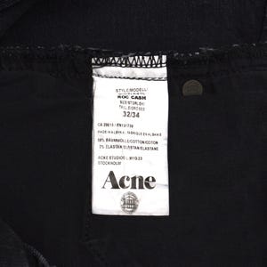 Pu&ograve; includere: Primo piano di jeans neri con un'etichetta bianca. L'etichetta recita "ROC CASH" e "32/34". L'etichetta include anche il marchio "Acne" e la localit&agrave; "Stoccolma". I jeans sono realizzati in cotone ed elastan.