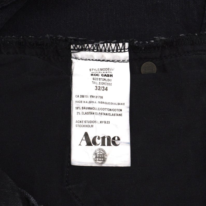 Pu&ograve; includere: Primo piano di jeans neri con un'etichetta bianca. L'etichetta recita "ROC CASH" e "32/34". L'etichetta include anche il marchio "Acne" e la localit&agrave; "Stoccolma". I jeans sono realizzati in cotone ed elastan.