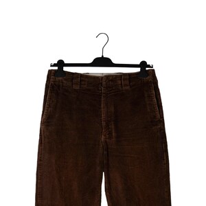 May include: Brown corduroy pants hanging on a black hanger. The pants have a classic five-pocket design and a straight leg. The corduroy fabric has a ribbed texture and a rich, warm brown color.