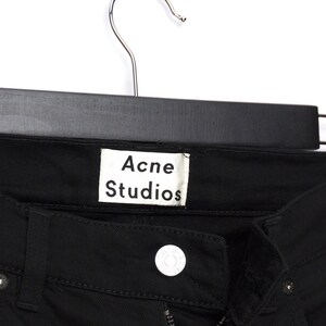 May include: Close-up of black denim jeans hanging on a black hanger. The white label reads "Acne Studios". The jeans have a button closure and a zipper. The fabric appears to be a medium weight.