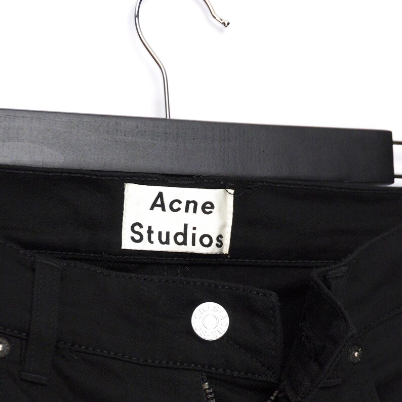May include: Close-up of black denim jeans hanging on a black hanger. The white label reads "Acne Studios". The jeans have a button closure and a zipper. The fabric appears to be a medium weight.