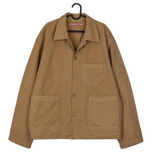 May include: A tan-colored jacket with a collared neckline, button-up front, and two large front pockets. The jacket has long sleeves and a small red tag on one pocket. The brand name "Acne Studios" is visible on a small tag near the collar.
