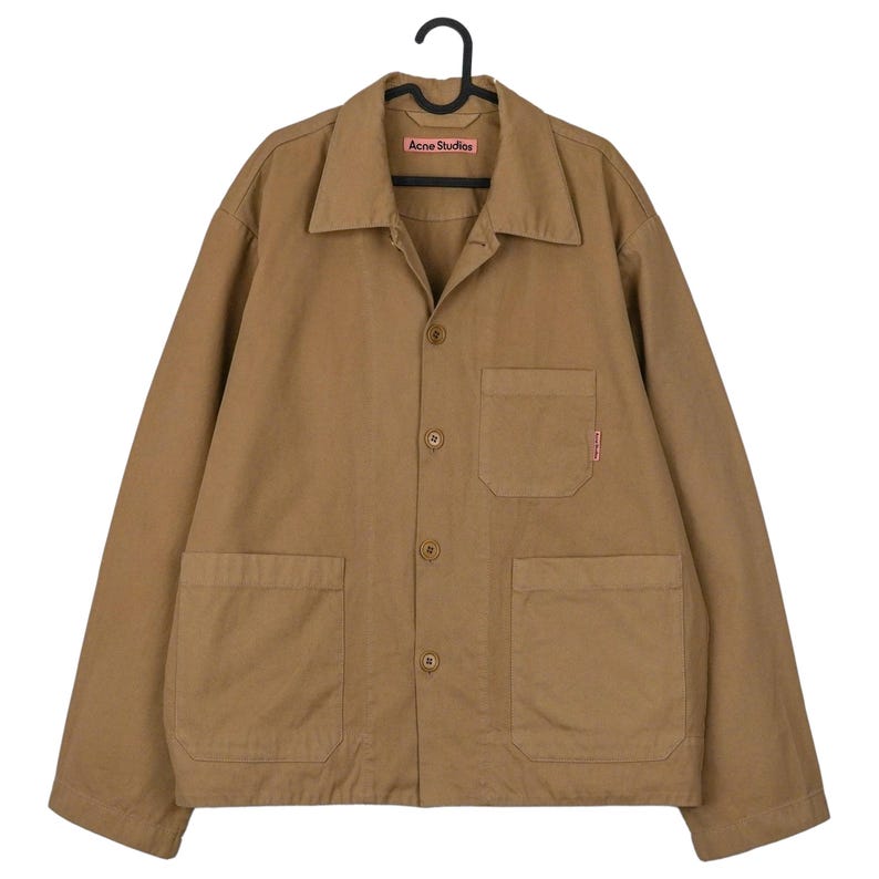 May include: A tan-colored jacket with a collared neckline, button-up front, and two large front pockets. The jacket has long sleeves and a small red tag on one pocket. The brand name "Acne Studios" is visible on a small tag near the collar.