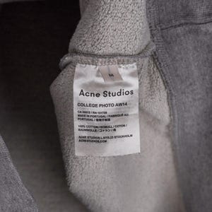 Puede incluir: Primer plano de una etiqueta de prenda gris de Acne Studios. La etiqueta blanca tiene texto negro, incluido el nombre de la marca, "COLLEGE PHOTO AW14", y detalles de la tela. La etiqueta est&aacute; adherida a una tela gris.