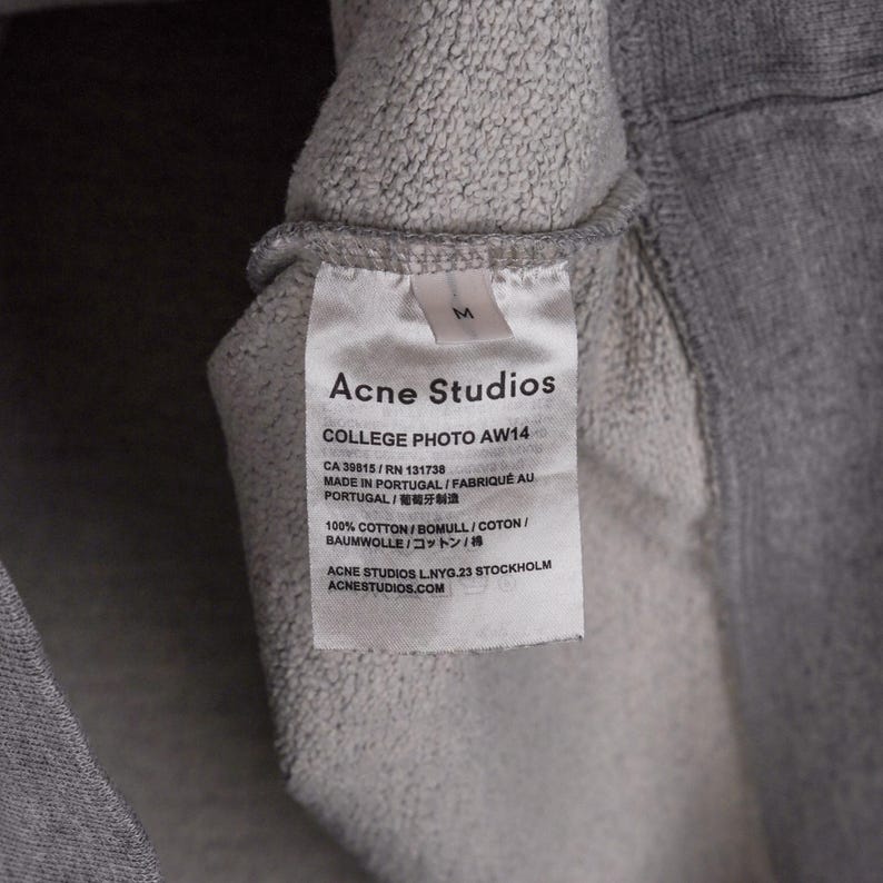 Puede incluir: Primer plano de una etiqueta de prenda gris de Acne Studios. La etiqueta blanca tiene texto negro, incluido el nombre de la marca, "COLLEGE PHOTO AW14", y detalles de la tela. La etiqueta est&aacute; adherida a una tela gris.