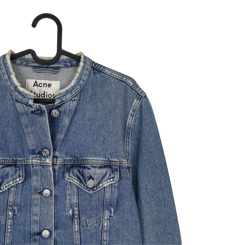 Op de afbeelding: Een spijkerjasje in een medium wassing met een gerafelde kraag en een zwarte hanger. De jas heeft een knoopsluiting, twee borstzakken en het label "Acne Studios". Klassieke stijl.