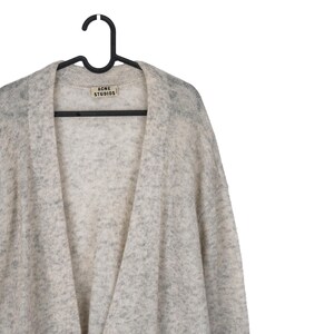 Puede incluir: Un c&aacute;rdigan gris claro con un patr&oacute;n moteado, colgado de una percha negra. La etiqueta dice "ACNE STUDIOS". El su&eacute;ter tiene un dise&ntilde;o frontal abierto y est&aacute; hecho de un material suave y texturizado.