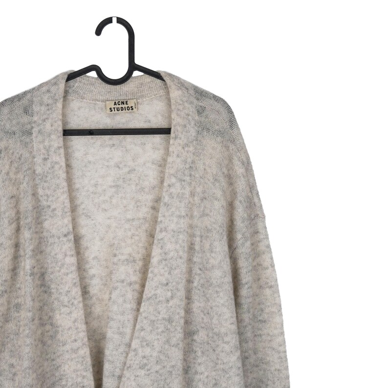 Puede incluir: Un c&aacute;rdigan gris claro con un patr&oacute;n moteado, colgado de una percha negra. La etiqueta dice "ACNE STUDIOS". El su&eacute;ter tiene un dise&ntilde;o frontal abierto y est&aacute; hecho de un material suave y texturizado.