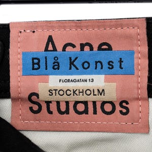 May include: A pink fabric label with the text "Acne Blå Konst" in black letters on a blue rectangle. Below, the label reads "FLORAGATAN 13 STOCKHOLM Studios". The label is stitched with white thread.