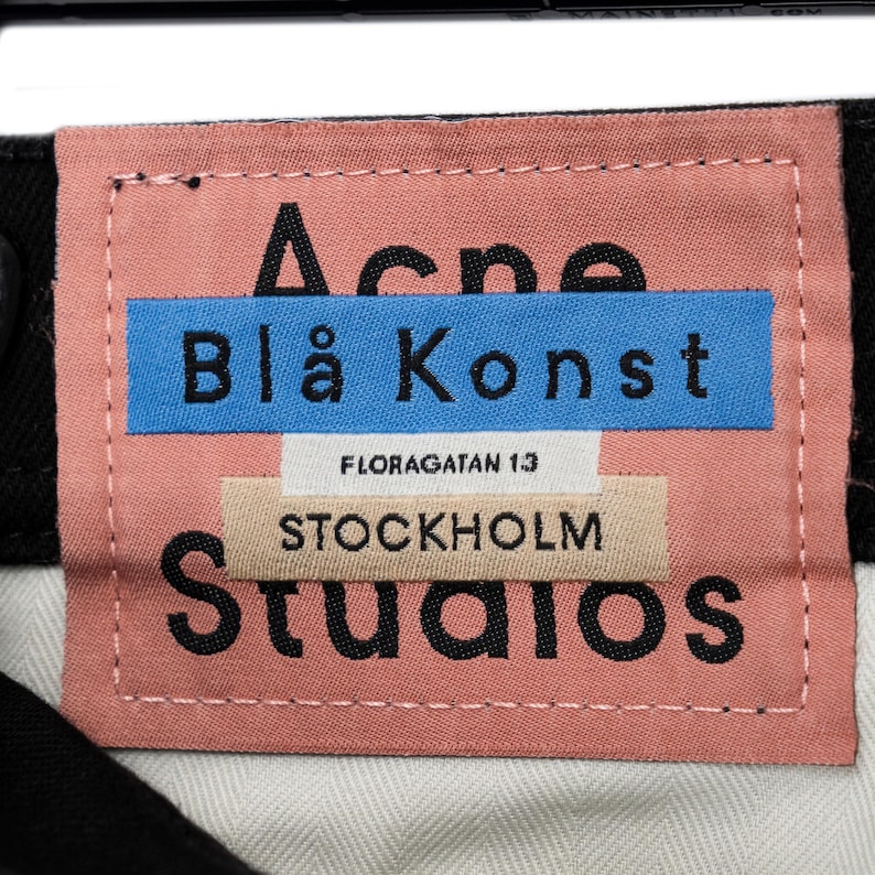 May include: A pink fabric label with the text "Acne Blå Konst" in black letters on a blue rectangle. Below, the label reads "FLORAGATAN 13 STOCKHOLM Studios". The label is stitched with white thread.