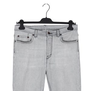 May include: Light gray denim jeans hanging on a black hanger. The jeans have a classic five-pocket design with a button and zipper closure. The denim appears to be a light wash, with subtle fading and stitching details.