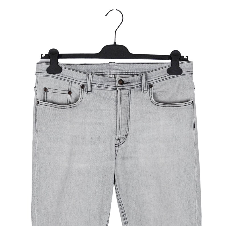May include: Light gray denim jeans hanging on a black hanger. The jeans have a classic five-pocket design with a button and zipper closure. The denim appears to be a light wash, with subtle fading and stitching details.