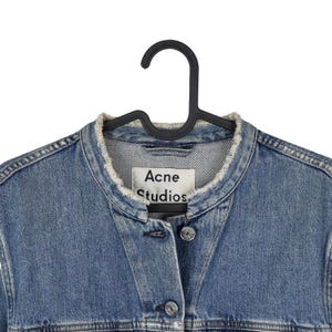 Op de afbeelding: Een blauw spijkerjasje met een gerafelde kraag en knopen. De jas hangt aan een zwarte hanger. Het label luidt "Acne Studios".
