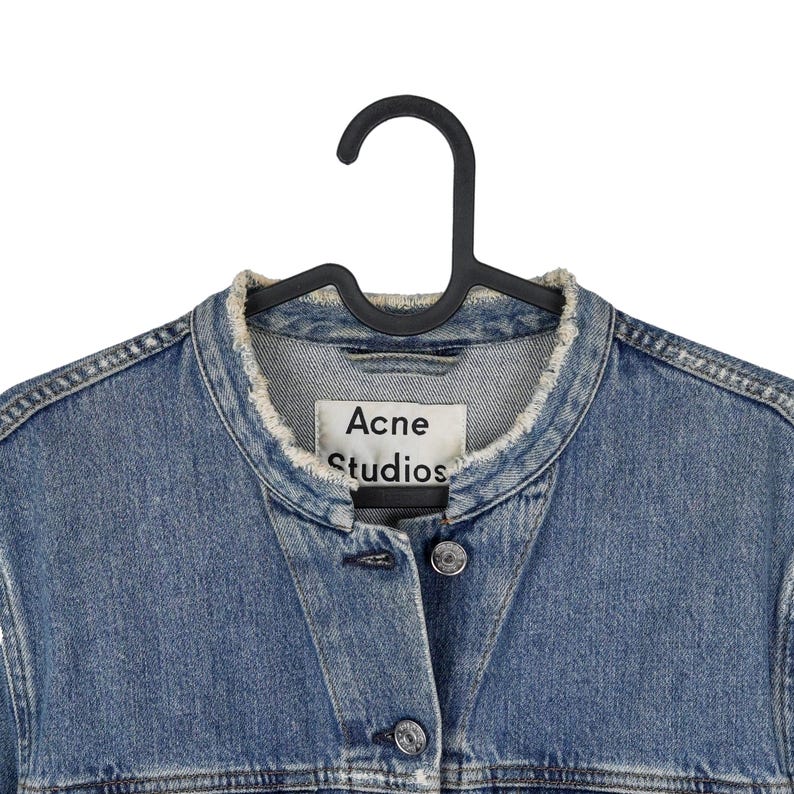 Op de afbeelding: Een blauw spijkerjasje met een gerafelde kraag en knopen. De jas hangt aan een zwarte hanger. Het label luidt "Acne Studios".