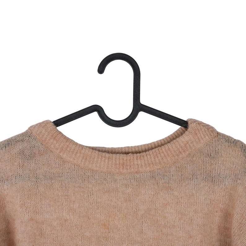Pu&ograve; includere: Un maglione color beige &egrave; appeso a una gruccia di plastica nera su sfondo bianco. Il maglione ha uno scollo rotondo e un aspetto leggermente trasparente. La gruccia &egrave; di design standard.
