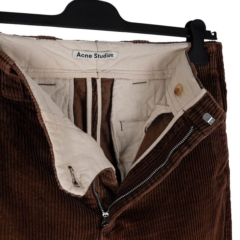 May include: Close-up of brown corduroy pants with a visible zipper and button closure. The pants are hanging on a black hanger. The brand name "Acne Studios" is visible on the inside waistband.