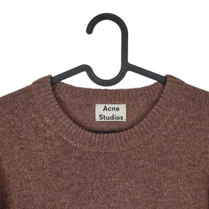 Peut inclure: Un pull marron &agrave; col rond de Acne Studios est suspendu &agrave; un cintre noir. Le pull a une encolure c&ocirc;tel&eacute;e et l'&eacute;tiquette de la marque est visible. Le tricot est de couleur unie et semble &ecirc;tre fait d'une mati&egrave;re douce.