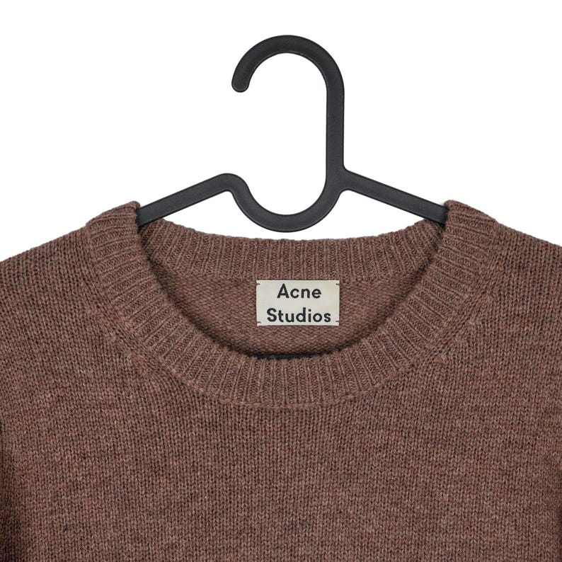 Peut inclure: Un pull marron &agrave; col rond de Acne Studios est suspendu &agrave; un cintre noir. Le pull a une encolure c&ocirc;tel&eacute;e et l'&eacute;tiquette de la marque est visible. Le tricot est de couleur unie et semble &ecirc;tre fait d'une mati&egrave;re douce.