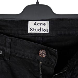 May include: Close-up of black denim jeans with the brand name "Acne Studios" on a white label. The jeans are hanging on a black hanger. A bronze-colored button is visible.
