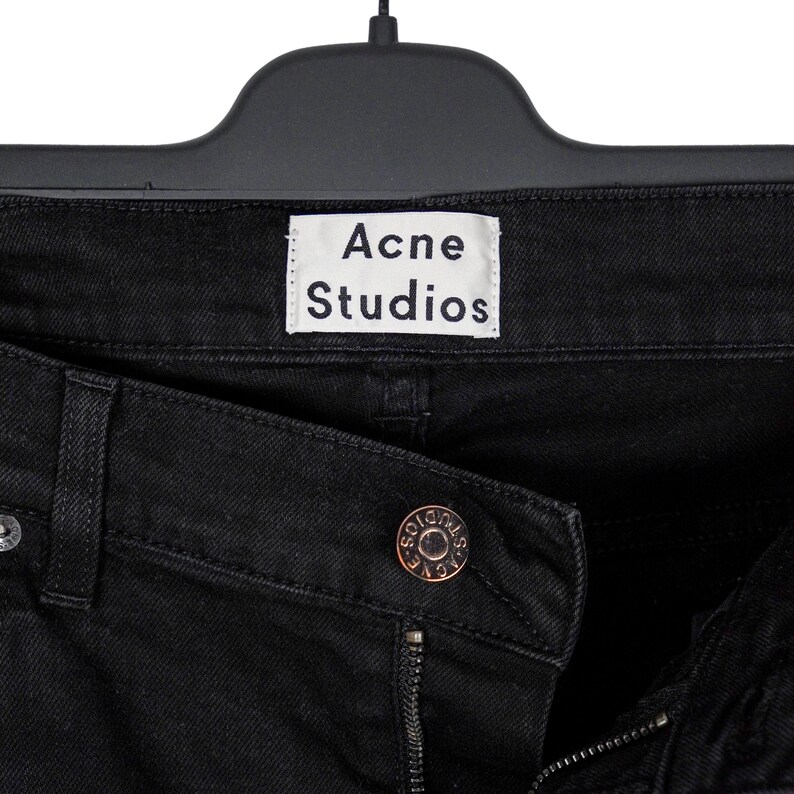 May include: Close-up of black denim jeans with the brand name "Acne Studios" on a white label. The jeans are hanging on a black hanger. A bronze-colored button is visible.