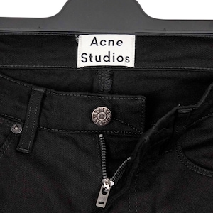 May include: Close-up of black denim jeans with a silver zipper and button. The jeans have a small white tag that reads "Acne Studios". The jeans are hanging on a black hanger.