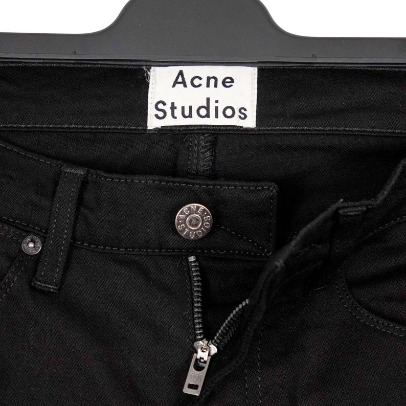 May include: Close-up of black denim jeans with a silver zipper and button. The jeans have a small white tag that reads "Acne Studios". The jeans are hanging on a black hanger.