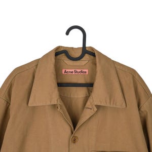 May include: A tan button-down shirt with a collar, hanging on a black hanger. The shirt has a small pink label that reads "Acne Studios". The shirt is made of a smooth fabric and has a single button closure.