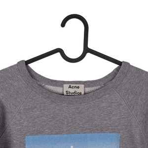 Puede incluir: Una sudadera gris de cuello redondo con la etiqueta "Acne Studios" visible. La sudadera est&aacute; colgada de una percha negra. La parte inferior de la sudadera tiene un gr&aacute;fico azul y blanco.
