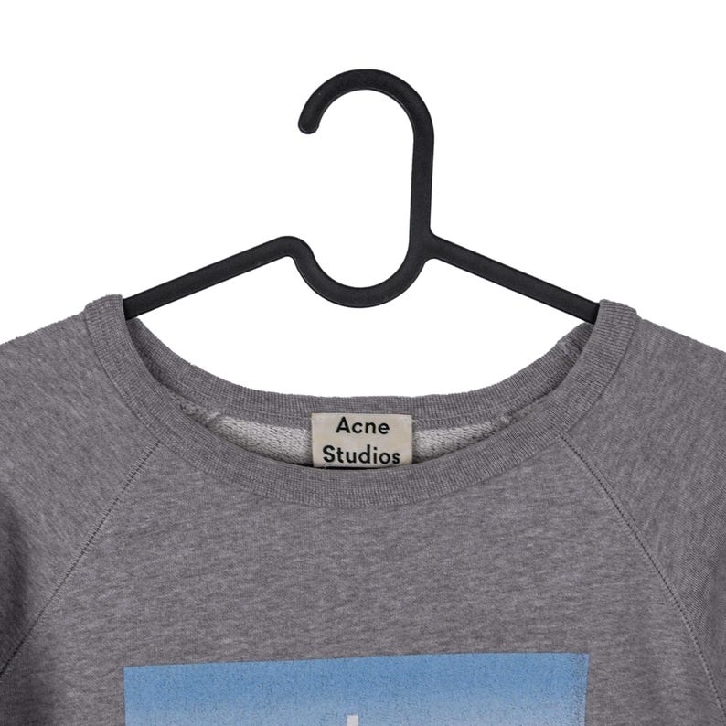 Puede incluir: Una sudadera gris de cuello redondo con la etiqueta "Acne Studios" visible. La sudadera est&aacute; colgada de una percha negra. La parte inferior de la sudadera tiene un gr&aacute;fico azul y blanco.