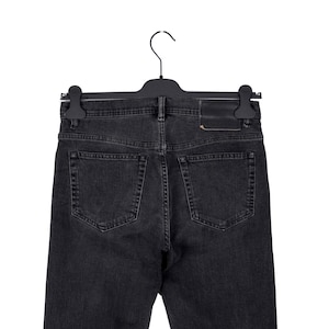 May include: Back view of black denim jeans hanging on a black hanger. The jeans have two back pockets and a leather patch. The denim is a dark gray color.