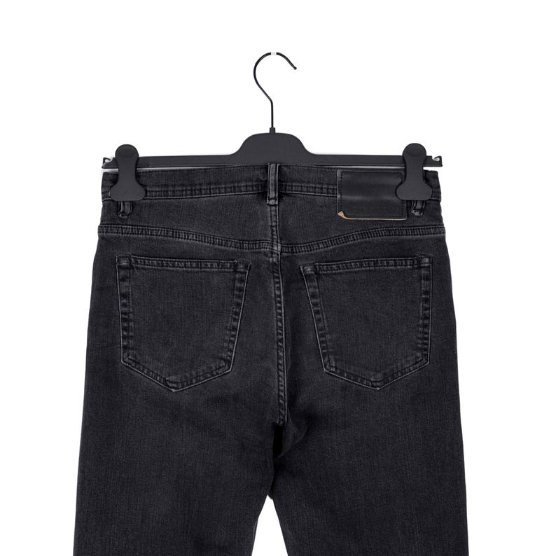 May include: Back view of black denim jeans hanging on a black hanger. The jeans have two back pockets and a leather patch. The denim is a dark gray color.