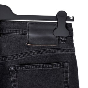 May include: Close-up of black denim jeans with a leather patch featuring the "Acne Studios" logo. The jeans are hanging on a black hanger. The denim has visible stitching and a back pocket.
