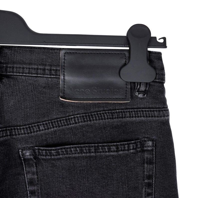 May include: Close-up of black denim jeans with a leather patch featuring the "Acne Studios" logo. The jeans are hanging on a black hanger. The denim has visible stitching and a back pocket.