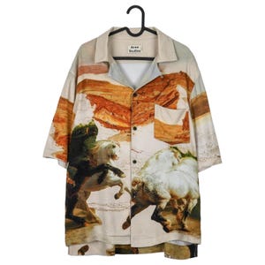 May include: Short-sleeved button-down shirt with a landscape and horse painting design. The shirt features a beige collar and the "Acne Studios" label. The artwork depicts a battle scene with horses and riders, in shades of brown, beige, and white.
