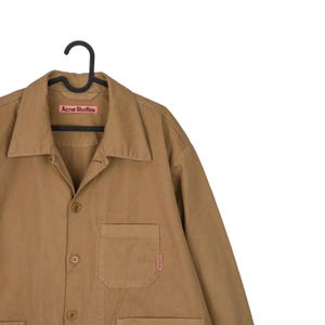 May include: A tan-colored button-up shirt with a collar and a single chest pocket. The shirt is hanging on a black hanger. The brand name "Acne Studios" is visible on a pink label.