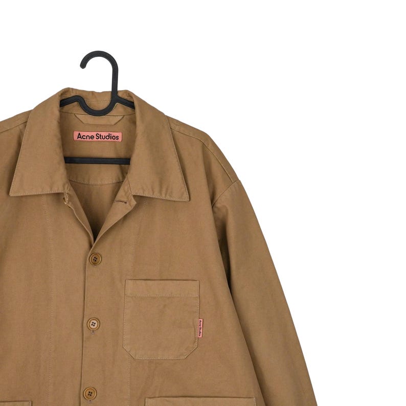 May include: A tan-colored button-up shirt with a collar and a single chest pocket. The shirt is hanging on a black hanger. The brand name "Acne Studios" is visible on a pink label.