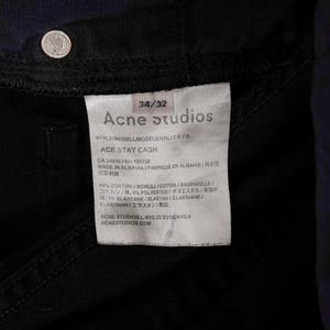May include: Close-up of a black denim garment with a white tag. The tag reads "Acne Studios" and "ACE STAY CASH". The garment has a silver button. The tag also lists the fabric composition and the location of manufacture.