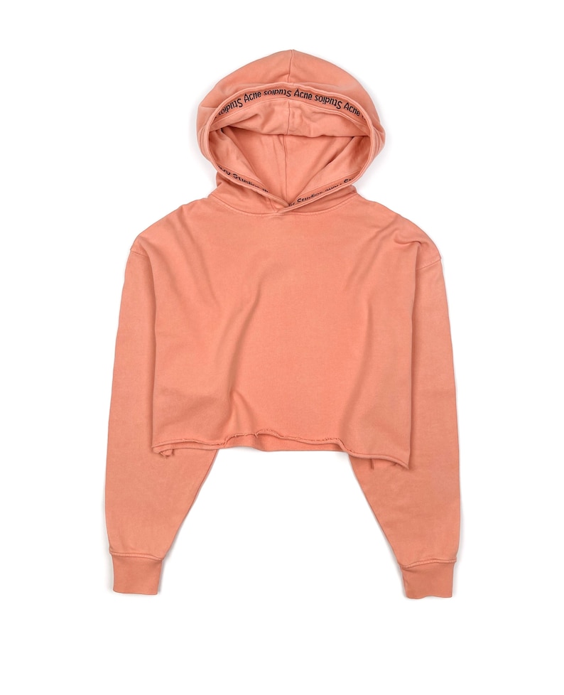 May include: A cropped, peach-colored hoodie with a drawstring hood. The hood features the text "Acne Studios" in black. The long sleeves have ribbed cuffs, and the bottom hem is raw-edged.