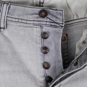 May include: Close-up of light gray denim jeans with a button fly. The jeans have a faded appearance and visible stitching. The buttons are a dark bronze color, and the brand name is visible on the top button.