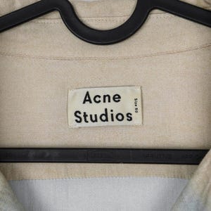 May include: Close-up of a beige shirt with the "Acne Studios" label. The label is rectangular with black text on a white background. The shirt is hanging on a black hanger. The size is 50.