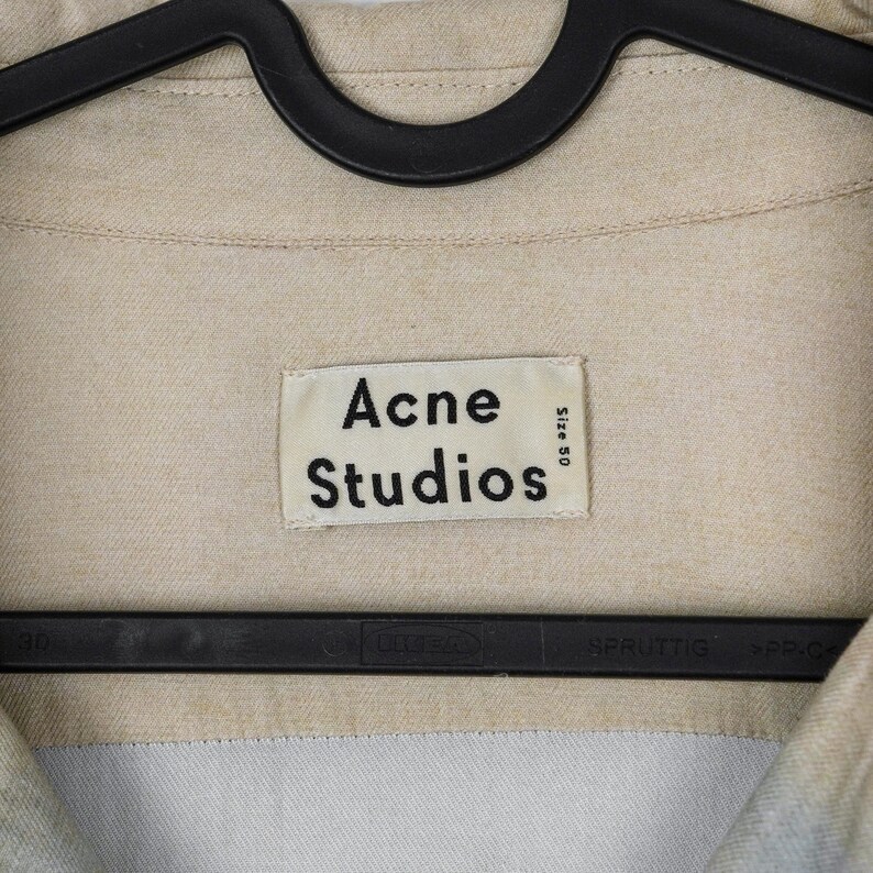 May include: Close-up of a beige shirt with the "Acne Studios" label. The label is rectangular with black text on a white background. The shirt is hanging on a black hanger. The size is 50.