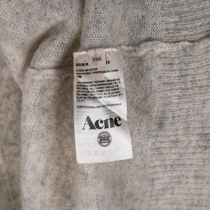 Puede incluir: Primer plano de una prenda de punto gris claro con una etiqueta. La etiqueta muestra la marca "Acne" y detalles de la composici&oacute;n de la tela, incluyendo mohair, lana, nailon y elastano. La prenda es de talla XXS.