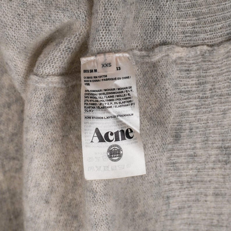 Puede incluir: Primer plano de una prenda de punto gris claro con una etiqueta. La etiqueta muestra la marca "Acne" y detalles de la composici&oacute;n de la tela, incluyendo mohair, lana, nailon y elastano. La prenda es de talla XXS.