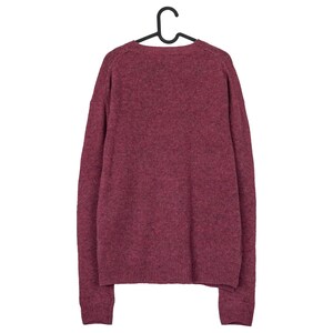 May include: A burgundy-coloured sweater hangs from a black hanger. The sweater has a crew neck and long sleeves. The knit fabric has a textured appearance. The sweater is a solid colour and appears to be made of a soft material.