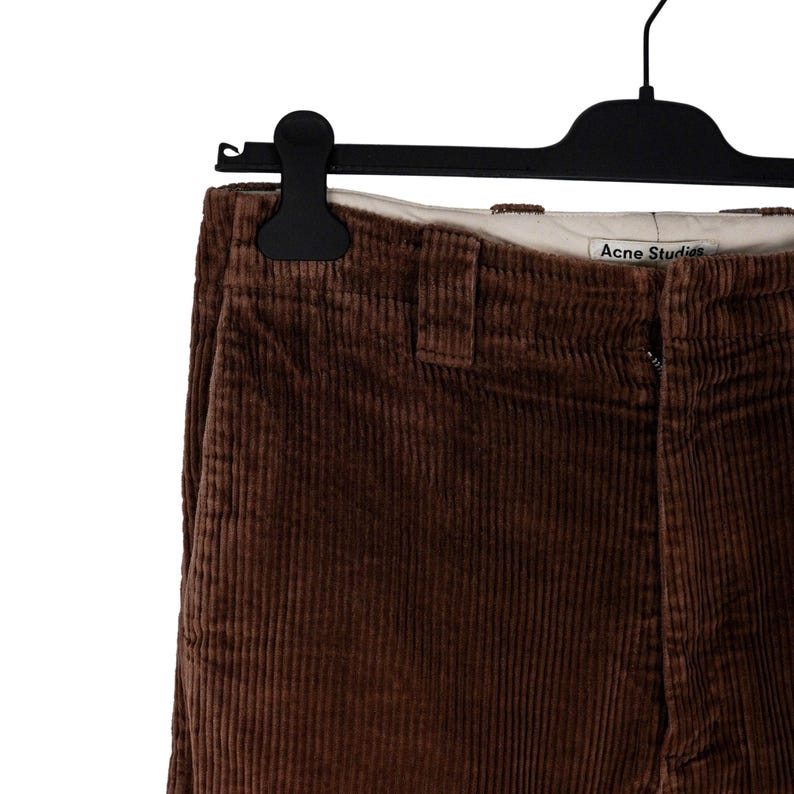 May include: Close-up of brown corduroy pants hanging on a black hanger. The pants have a vertical ribbed texture and a visible zipper. The brand name "Acne Studios" is visible on the waistband.