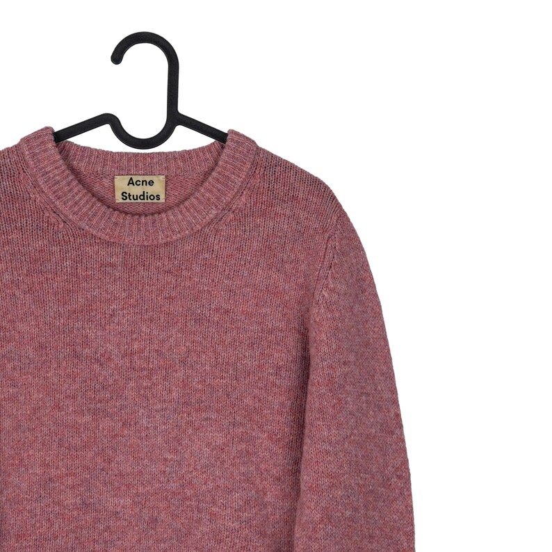 May include: A dusty rose-colored sweater with a crew neck, hanging on a black hanger. The label reads "Acne Studios". The sweater has a textured knit appearance.