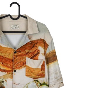 May include: A short-sleeved shirt with a landscape painting design. The shirt features a button-down front, a chest pocket, and a collar. The tag reads "Acne Studios". The shirt is hung on a black hanger.