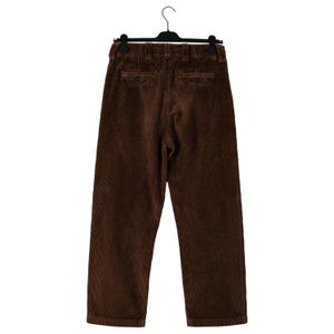 May include: Brown corduroy pants hanging on a black hanger. The pants have a classic design with a button and zipper closure, and two back pockets. The corduroy fabric has a vertical ribbed texture.